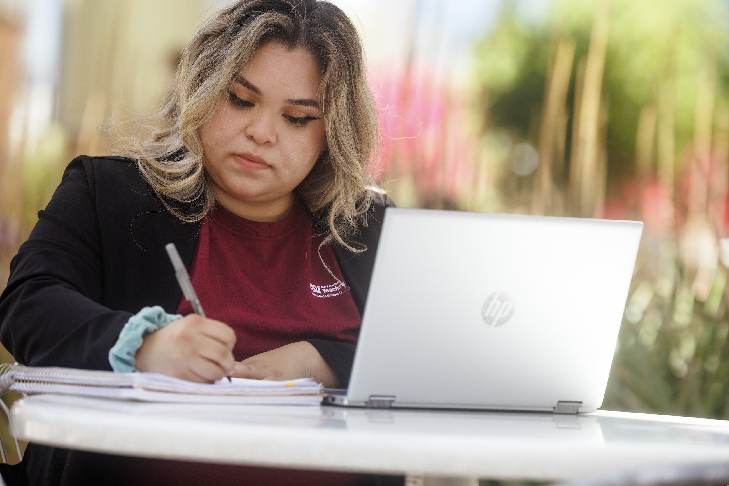 Student writing in a notebook with a laptop on an outside table